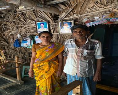 A person and person standing in front of a straw hut

Description automatically generated