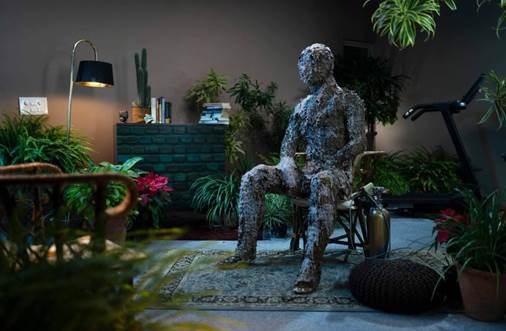 A statue of a person sitting in a chair in a room with plants

Description automatically generated